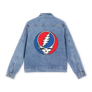 Puede incluir: Una chaqueta de mezclilla azul claro con un gráfico circular grande en la parte posterior. El diseño muestra una calavera estilizada con un rayo blanco sobre un fondo rojo y azul, centrado en el panel trasero de la prenda de vestir.