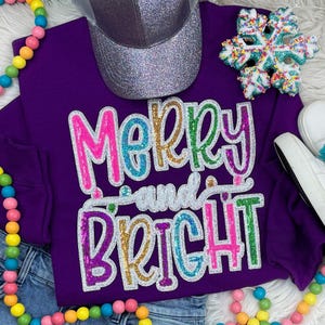 May include: Purple sweatshirt with the text "Merry and Bright" in colorful sequins. The sweatshirt is laid out on a white surface with a silver glitter trucker hat, a snowflake, and a beaded necklace.