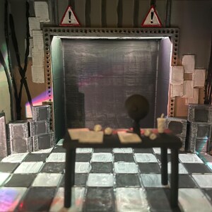 May include: A miniature diorama of a room with a black and white chequered floor, a central black table with a small globe and papers, stacks of grey boxes, and two red triangular warning signs with exclamation marks mounted above a dark doorway.