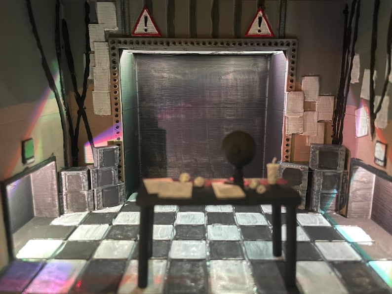 May include: A miniature diorama of a room with a black and white chequered floor, a central black table with a small globe and papers, stacks of grey boxes, and two red triangular warning signs with exclamation marks mounted above a dark doorway.