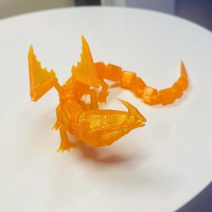 Puede incluir: Figura de escorpión naranja impresa en 3D. El escorpión tiene un cuerpo segmentado, una cola curvada y dos grandes pinzas. La figura está hecha de plástico naranja translúcido y está sobre una superficie blanca.