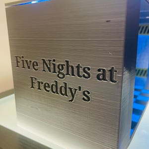 K&ouml;nnte beinhalten: Eine rechteckige, silberfarbene 3D-gedruckte Display-Box mit dem eingepr&auml;gten Text "Five Nights at Freddy's" auf der Vorderseite. Das Innere der Box ist hellblau beleuchtet und zeigt eine abgestufte Struktur im Inneren.