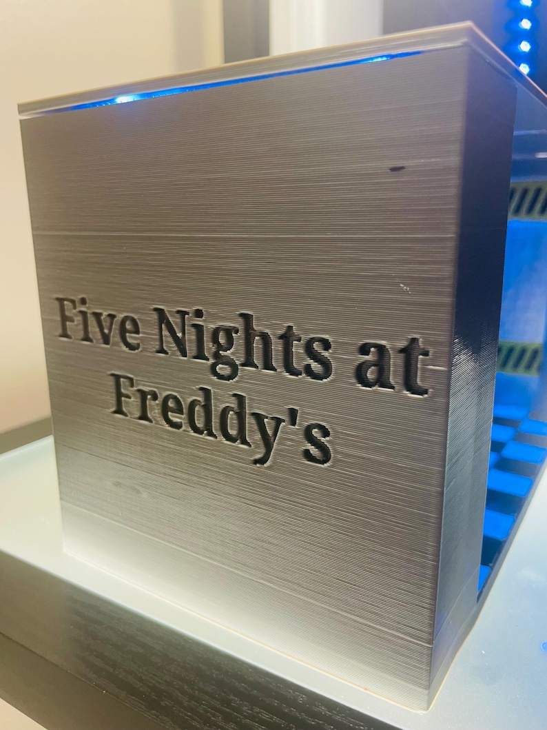 K&ouml;nnte beinhalten: Eine rechteckige, silberfarbene 3D-gedruckte Display-Box mit dem eingepr&auml;gten Text "Five Nights at Freddy's" auf der Vorderseite. Das Innere der Box ist hellblau beleuchtet und zeigt eine abgestufte Struktur im Inneren.