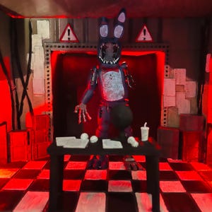 May include: A diorama featuring a dark blue, weathered rabbit figure with a missing face and exposed mechanical parts, standing behind a black table with white papers and a cup, set in a room with a red-lit checkered floor and warning signs.