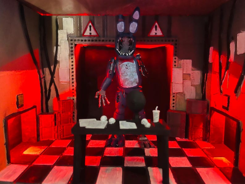 May include: A diorama featuring a dark blue, weathered rabbit figure with a missing face and exposed mechanical parts, standing behind a black table with white papers and a cup, set in a room with a red-lit checkered floor and warning signs.