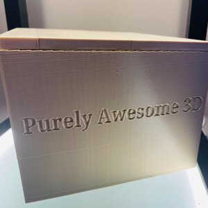 May include: A beige, rectangular 3D printed box with a flat top and a smooth surface. The front face of the box features the text "Purely Awesome 3D" embossed in a clear, sans-serif font. The object displays visible horizontal layer lines.