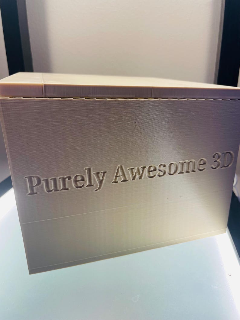 May include: A beige, rectangular 3D printed box with a flat top and a smooth surface. The front face of the box features the text "Purely Awesome 3D" embossed in a clear, sans-serif font. The object displays visible horizontal layer lines.