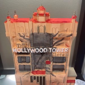 Op de afbeelding: Een 3D-geprint model van het Hollywood Tower Hotel, met een perzikgekleurde gevel en rode accenten op het dak. De tekst "The Hollywood Tower Hotel" staat in witte letters.