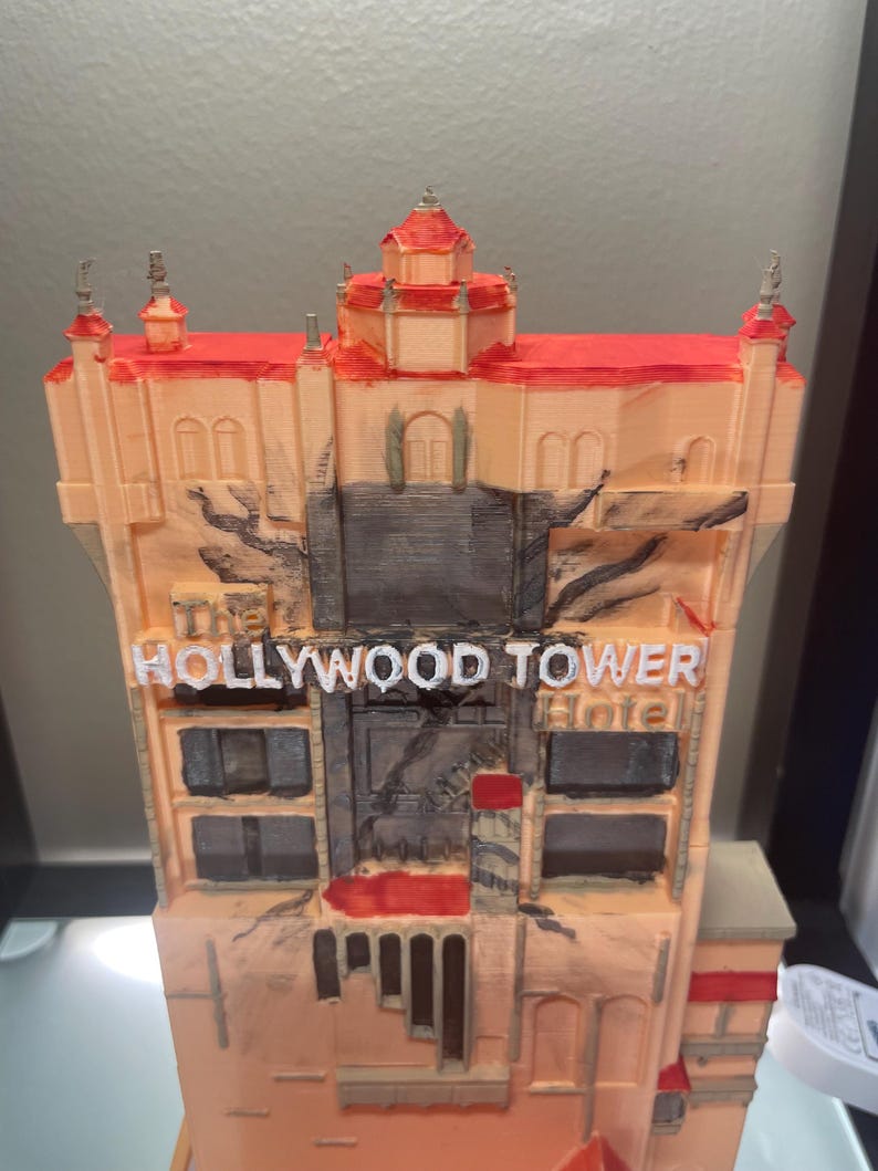 Op de afbeelding: Een 3D-geprint model van het Hollywood Tower Hotel, met een perzikgekleurde gevel en rode accenten op het dak. De tekst "The Hollywood Tower Hotel" staat in witte letters.