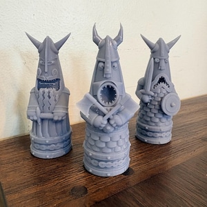 Op de afbeelding: Drie grijze, 3D-geprinte beeldjes die gestileerde Vikingkrijgers voorstellen. Elk figuur draagt een helm met hoorns en heeft een wijd open mond met zichtbare tanden. De figuren dragen gestructureerde harnassen en houden bijlen en zwaarden vast.
