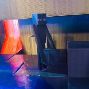 Puede incluir: Una figura de personaje negro de Minecraft de pie en una plataforma con un fondo azul y rojo. La figura est&aacute; hecha de pl&aacute;stico y tiene un dise&ntilde;o pixelado.