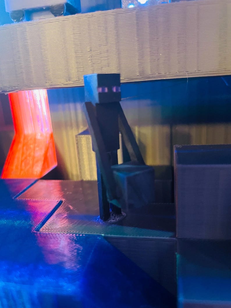 Puede incluir: Una figura de personaje negro de Minecraft de pie en una plataforma con un fondo azul y rojo. La figura est&aacute; hecha de pl&aacute;stico y tiene un dise&ntilde;o pixelado.