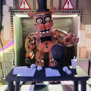 May include: A brown bear animatronic figure with a black top hat and bow tie stands behind a black desk. The desk holds white papers, crumpled paper balls, a black circular object, and a white cup. The background features a chequered floor and walls with two red and white warning signs with exclamation marks.