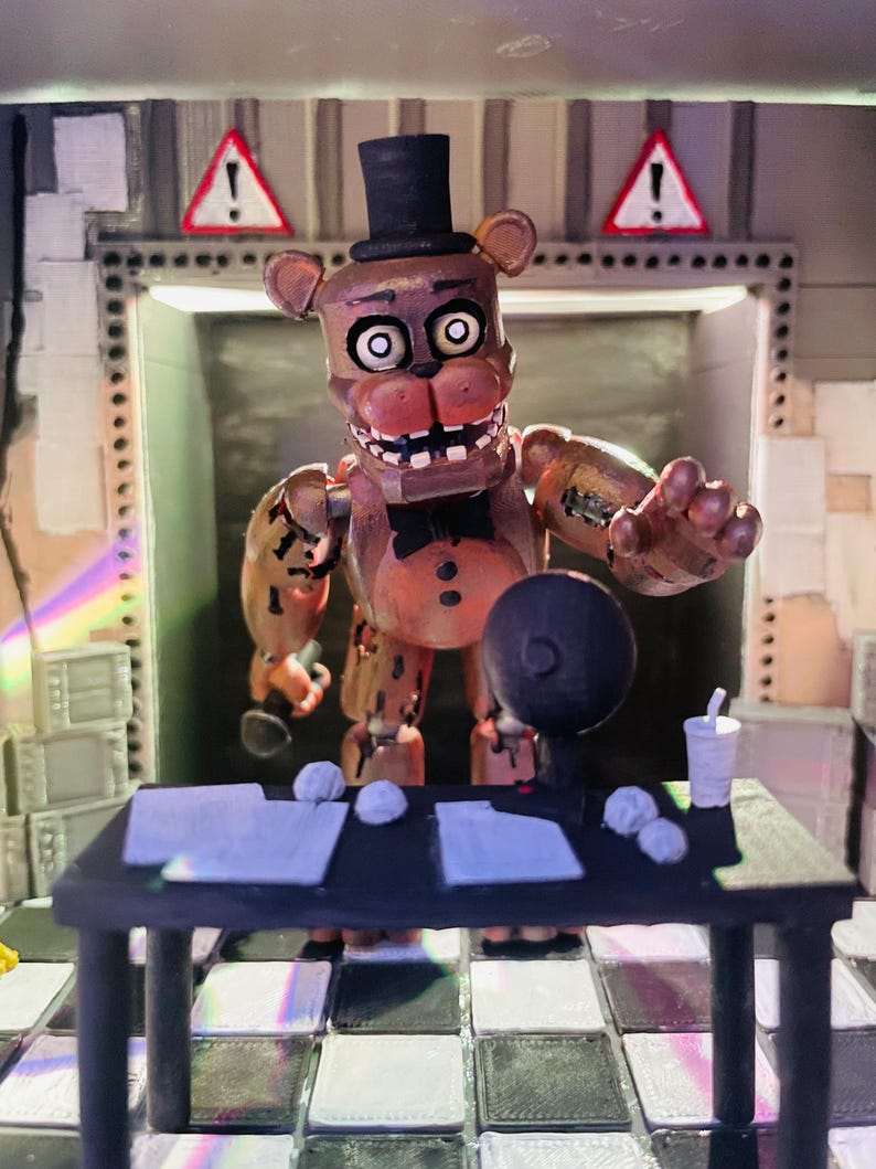 May include: A brown bear animatronic figure with a black top hat and bow tie stands behind a black desk. The desk holds white papers, crumpled paper balls, a black circular object, and a white cup. The background features a chequered floor and walls with two red and white warning signs with exclamation marks.