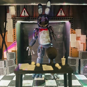 May include: A diorama featuring a weathered, blue animatronic rabbit figure with exposed mechanical parts, a red bowtie, and a damaged face. The figure stands in a dark room with a black desk, papers, a small cup, and warning signs on the wall.