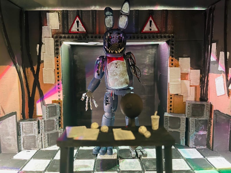 May include: A diorama featuring a weathered, blue animatronic rabbit figure with exposed mechanical parts, a red bowtie, and a damaged face. The figure stands in a dark room with a black desk, papers, a small cup, and warning signs on the wall.