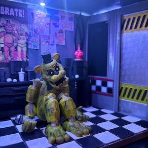 K&ouml;nnte beinhalten: Eine goldene B&auml;ren-Animatronic-Figur mit schwarzem Zylinder und Fliege sitzt auf einem schwarz-wei&szlig; karierten Boden. Die Szene enth&auml;lt ein Wandplakat mit dem Text CELEBRATE!, verschiedene Zeichnungen, einen kleinen rosa Cupcake und eine gelb gestreifte T&uuml;r.