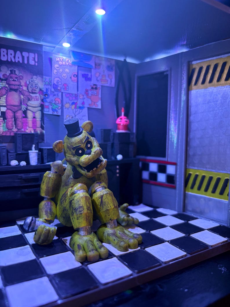 K&ouml;nnte beinhalten: Eine goldene B&auml;ren-Animatronic-Figur mit schwarzem Zylinder und Fliege sitzt auf einem schwarz-wei&szlig; karierten Boden. Die Szene enth&auml;lt ein Wandplakat mit dem Text CELEBRATE!, verschiedene Zeichnungen, einen kleinen rosa Cupcake und eine gelb gestreifte T&uuml;r.