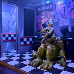 K&ouml;nnte beinhalten: Eine goldene B&auml;ren-Animatronic-Figur mit einem schwarzen Zylinder sitzt auf einem schwarz-wei&szlig; karierten Boden. Die Szene enth&auml;lt ein Wandplakat mit dem Text CELEBRATE! und verschiedene Kinderzeichnungen. Die Beleuchtung ist gedimmt mit Blaut&ouml;nen.