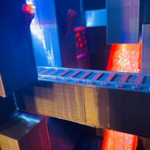 Puede incluir: Un modelo 3D impreso de una estructura futurista con un flujo de lava roja incandescente y una pasarela iluminada en azul. El modelo est&aacute; hecho de pl&aacute;stico y tiene una superficie texturizada.