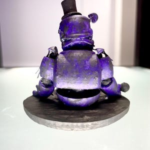 Puede incluir: Figura morada de un oso con sombrero de copa negro, sobre una base circular gris oscuro. La figura tiene detalles mec&aacute;nicos visibles y un aspecto desgastado. El fondo es blanco suave. El dise&ntilde;o sugiere un estilo vintage o estilizado.