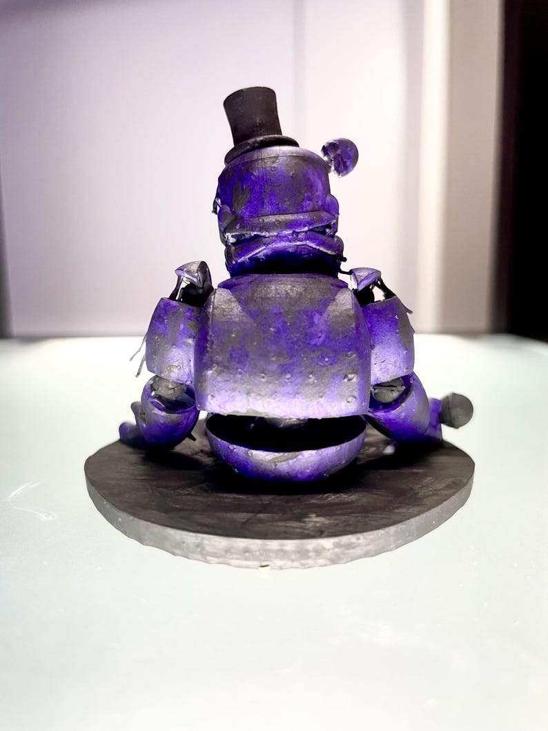 Puede incluir: Figura morada de un oso con sombrero de copa negro, sobre una base circular gris oscuro. La figura tiene detalles mec&aacute;nicos visibles y un aspecto desgastado. El fondo es blanco suave. El dise&ntilde;o sugiere un estilo vintage o estilizado.