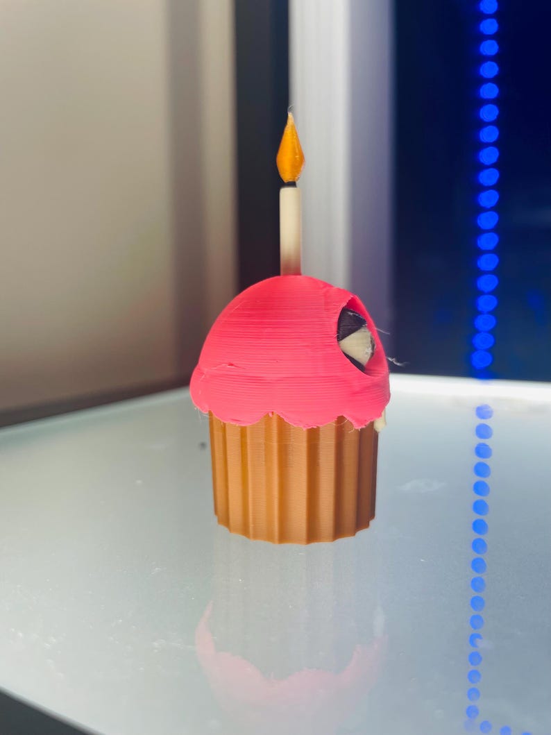 May include: A 3D printed cupcake figurine featuring a brown fluted base and a bright pink frosting top. A single candle with a yellow flame sits centred on the frosting. A small black and white circular eye detail is visible on the side of the pink frosting.