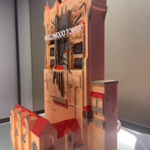 Op de afbeelding: Een 3D-geprint model van het Hollywood Tower Hotel, met een perzikkleurige buitenkant en rode dakaccenten. Het model heeft gedetailleerde architectonische kenmerken, waaronder ramen en de naam van het hotel. Het is een miniatuurreplica van het iconische gebouw.