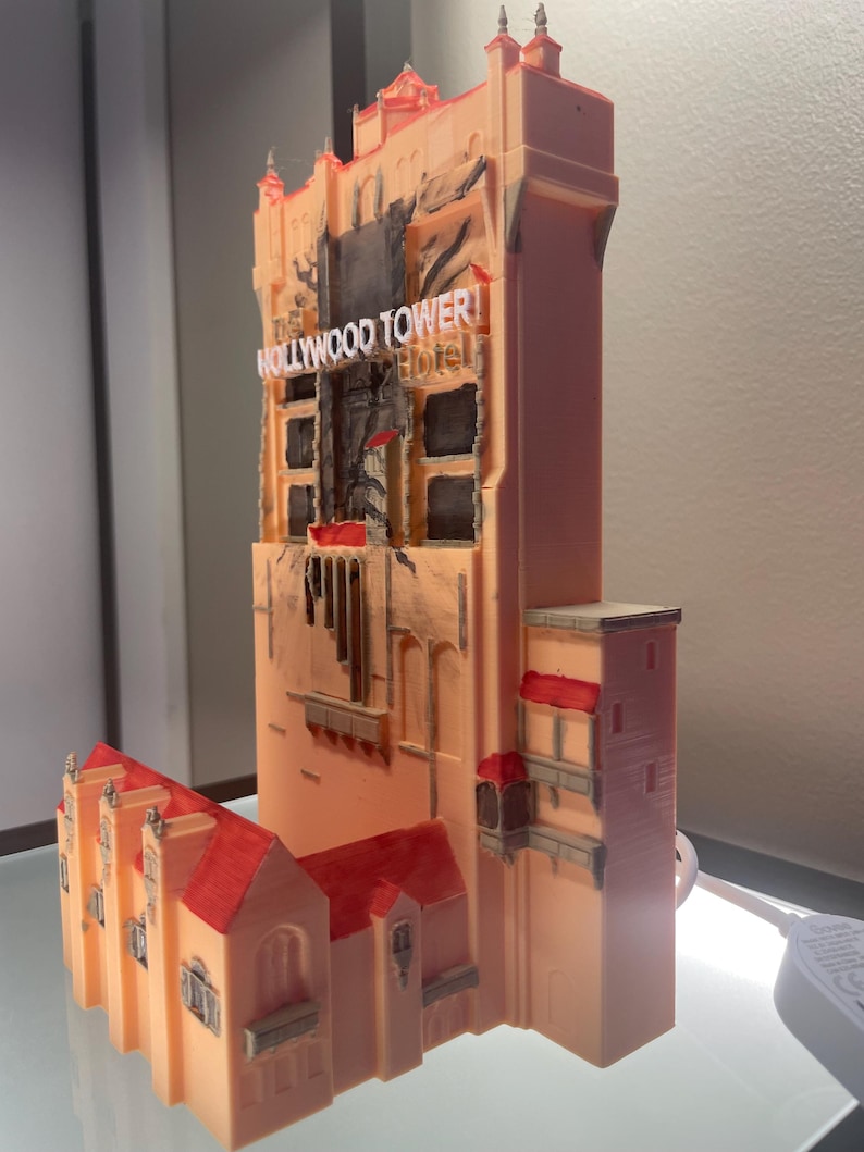Op de afbeelding: Een 3D-geprint model van het Hollywood Tower Hotel, met een perzikkleurige buitenkant en rode dakaccenten. Het model heeft gedetailleerde architectonische kenmerken, waaronder ramen en de naam van het hotel. Het is een miniatuurreplica van het iconische gebouw.