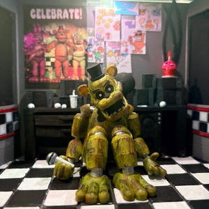 K&ouml;nnte beinhalten: Eine goldene B&auml;ren-Animatronic-Figur mit schwarzem Zylinder und Fliege, die ein kleines silbernes Mikrofon h&auml;lt. Die Figur sitzt auf einem schwarz-wei&szlig; karierten Boden vor einer Wand, die mit einem CELEBRATE!-Poster und verschiedenen Zeichnungen verziert ist.