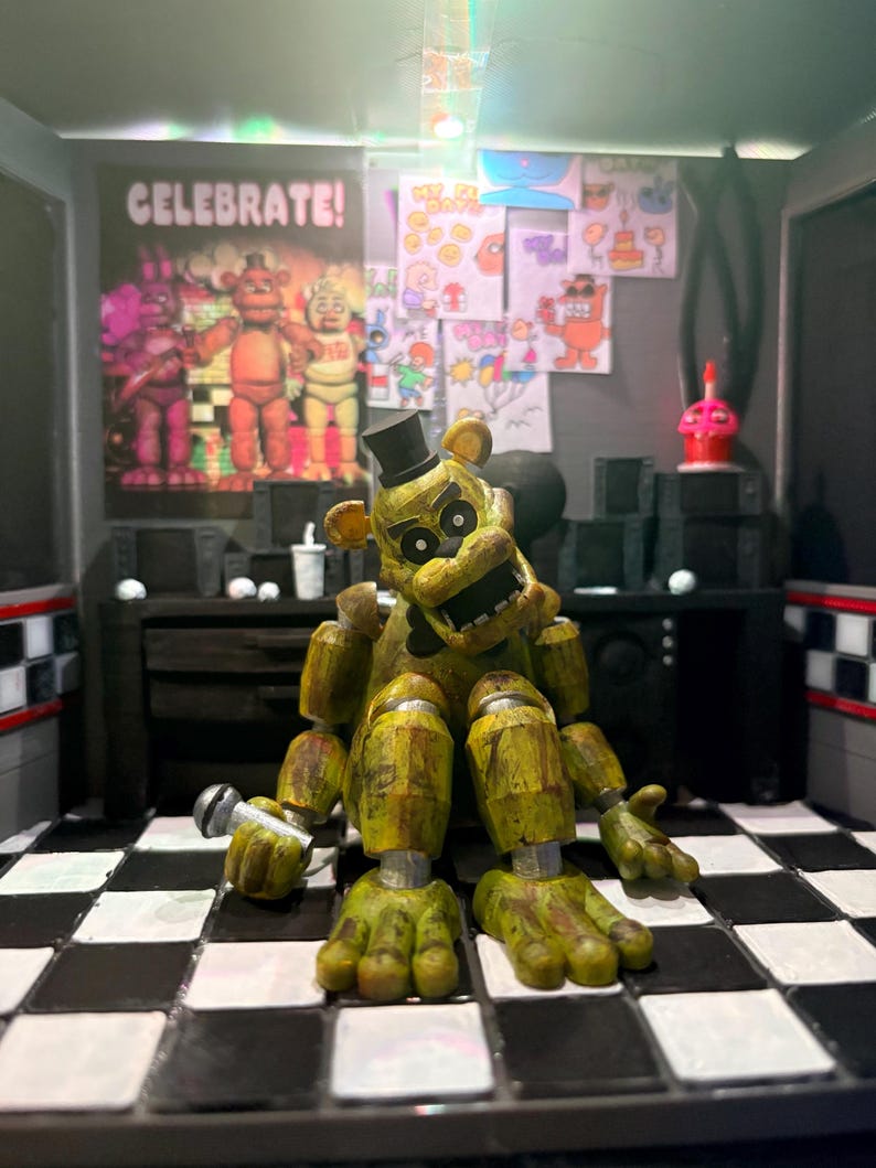 K&ouml;nnte beinhalten: Eine goldene B&auml;ren-Animatronic-Figur mit schwarzem Zylinder und Fliege, die ein kleines silbernes Mikrofon h&auml;lt. Die Figur sitzt auf einem schwarz-wei&szlig; karierten Boden vor einer Wand, die mit einem CELEBRATE!-Poster und verschiedenen Zeichnungen verziert ist.