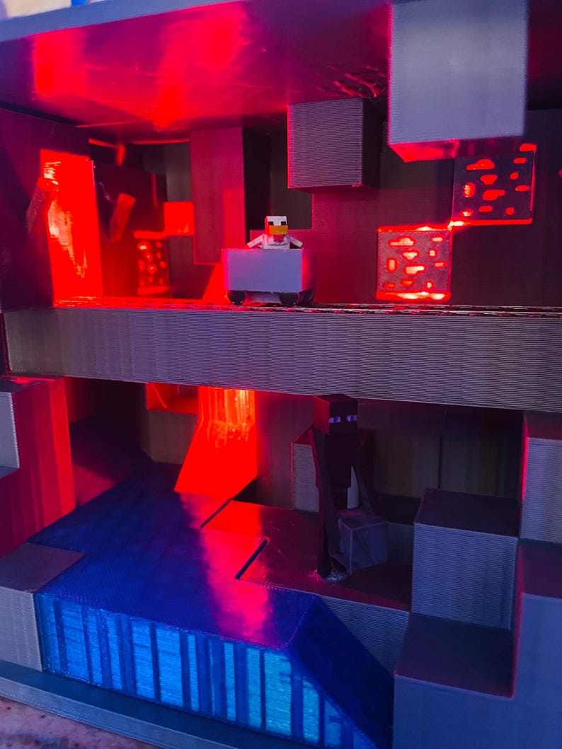 Puede incluir: Un diorama impreso en 3D con tem&aacute;tica de Minecraft con un esquema de iluminaci&oacute;n rojo y azul. Una peque&ntilde;a figura de un pollo montando un carro de minas est&aacute; en el nivel superior. Una figura de un personaje de piel negra con ojos morados est&aacute; en el nivel inferior.