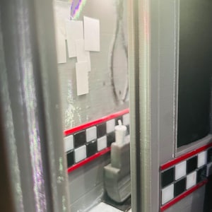 K&ouml;nnte beinhalten: Ein Miniatur-Diorama mit einem grauen Innenraum, einer schwarz-wei&szlig; karierten Bord&uuml;re und einer roten Zierlinie. Die Szene enth&auml;lt einen kleinen grauen Stuhl mit einem wei&szlig;en Becher darauf, wei&szlig;e rechteckige Wandakzente und einen Boden.