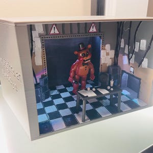 May include: A 3D-printed diorama box featuring a brown bear character with a black top hat and bowtie standing in a room with a black and white chequered floor. The scene includes a black table, warning signs, and hanging wires against a grey wall.
