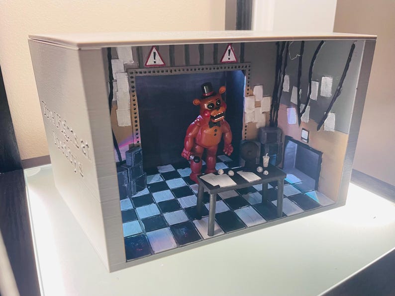 May include: A 3D-printed diorama box featuring a brown bear character with a black top hat and bowtie standing in a room with a black and white chequered floor. The scene includes a black table, warning signs, and hanging wires against a grey wall.