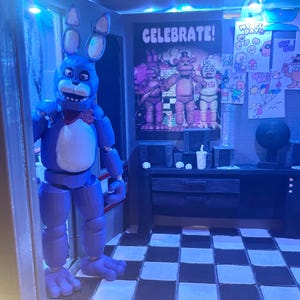 K&ouml;nnte beinhalten: Eine blaue Animatronic-Hasenfigur mit wei&szlig;er Brust und roter Fliege steht in einem T&uuml;rrahmen. Dahinter befindet sich ein Raum mit schwarz-wei&szlig; kariertem Boden, einem Schreibtisch mit Becher und einem Poster mit dem Text CELEBRATE!.