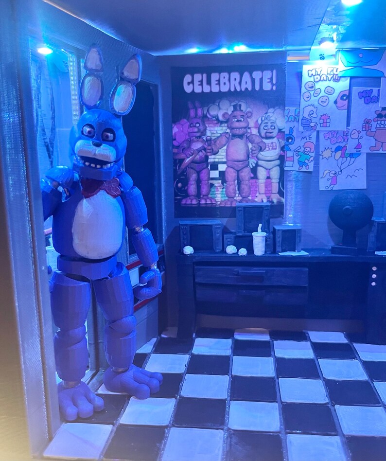 K&ouml;nnte beinhalten: Eine blaue Animatronic-Hasenfigur mit wei&szlig;er Brust und roter Fliege steht in einem T&uuml;rrahmen. Dahinter befindet sich ein Raum mit schwarz-wei&szlig; kariertem Boden, einem Schreibtisch mit Becher und einem Poster mit dem Text CELEBRATE!.