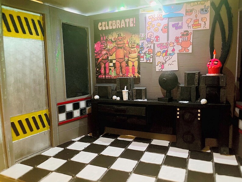 K&ouml;nnte beinhalten: Ein Miniatur-Diorama eines Sicherheitsb&uuml;ros mit schwarz-wei&szlig; kariertem Boden, einem Schreibtisch mit elektronischen Ger&auml;ten, einem Celebrate-Poster mit drei Figuren, Kinderzeichnungen an der Wand und einer T&uuml;r mit gelben Warnstreifen.