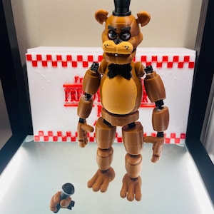 May include: A brown articulated bear figure with a black top hat and bow tie, holding a microphone. The figure is standing in front of a white box with a red and white checkered pattern and text. The figure is a toy.