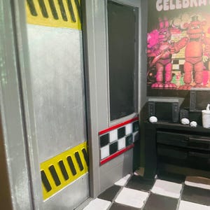 K&ouml;nnte beinhalten: Ein Miniatur-Diorama mit einer silbernen T&uuml;r mit gelb-schwarzen Warnstreifen, einem schwarz-wei&szlig; karierten Wandakzent und einem schwarz-wei&szlig; gefliesten Boden. Ein Poster zeigt zwei Zeichentrickfiguren mit dem Text CELEBRATE.