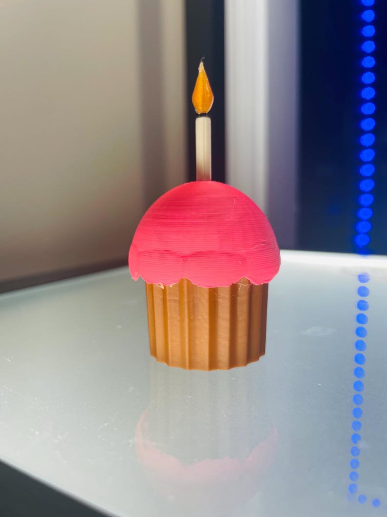 May include: A 3D printed cupcake figure featuring a tan fluted base and a bright pink rounded top. A white candle with a translucent orange flame sits upright in the centre of the pink frosting, creating a decorative object with a smooth finish.