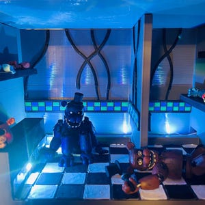 Op de afbeelding: Een diorama met Five Nights at Freddy's-figuren in een kamer met een zwart-wit geblokte vloer, blauwe muren en groen-blauwe tegelaccenten. De sc&egrave;ne bevat een donkerblauw berenfiguur, een bruin berenfiguur en een gele vogelkop.