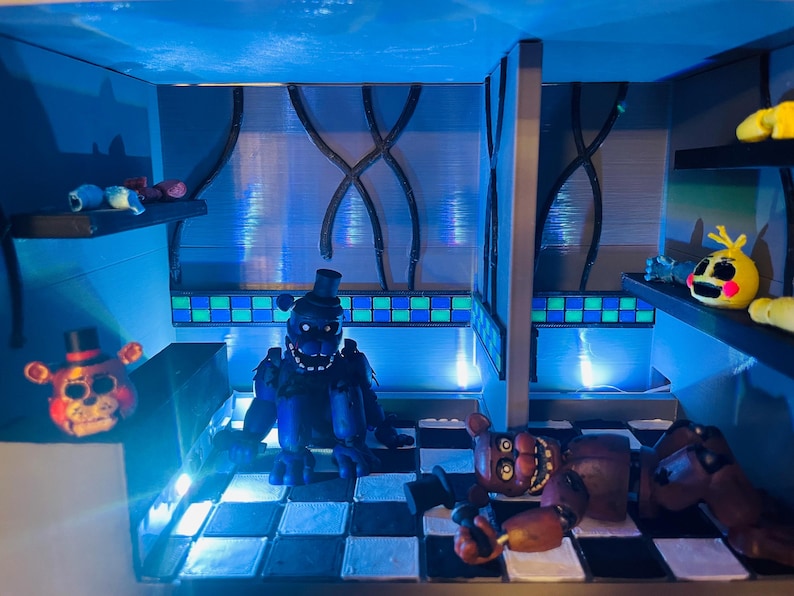 Op de afbeelding: Een diorama met Five Nights at Freddy's-figuren in een kamer met een zwart-wit geblokte vloer, blauwe muren en groen-blauwe tegelaccenten. De sc&egrave;ne bevat een donkerblauw berenfiguur, een bruin berenfiguur en een gele vogelkop.