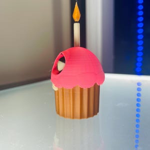 May include: A 3D printed cupcake figurine featuring a brown fluted base and a bright pink frosting top. A single white candle with a yellow flame sits upright in the centre of the frosting. A small dark opening is visible on the side of the pink top.