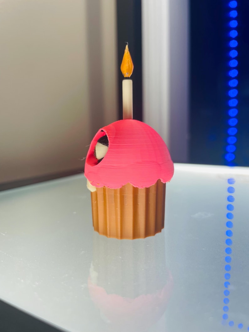 May include: A 3D printed cupcake figurine featuring a brown fluted base and a bright pink frosting top. A single white candle with a yellow flame sits upright in the centre of the frosting. A small dark opening is visible on the side of the pink top.