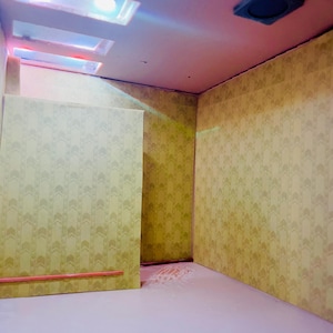 May include: A miniature room diorama featuring yellow wallpaper with a repeating chevron pattern, a light-colored floor, and a ceiling with three rectangular recessed light fixtures and one square dark vent cover, illuminated by soft pink and blue lighting.