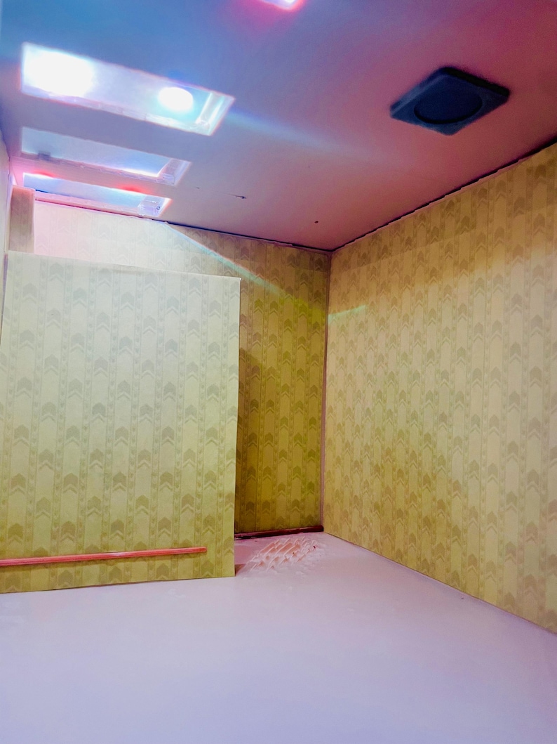 May include: A miniature room diorama featuring yellow wallpaper with a repeating chevron pattern, a light-colored floor, and a ceiling with three rectangular recessed light fixtures and one square dark vent cover, illuminated by soft pink and blue lighting.