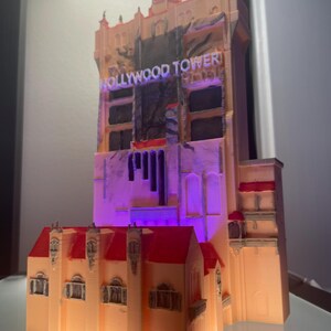 Op de afbeelding: Een 3D-geprint model van het Hollywood Tower Hotel, met een beige buitenkant en rode dakdetails. Het model heeft de verlichte tekst "HOLLYWOOD TOWER". Het gebouw wordt verlicht met paarse en oranje lichten.