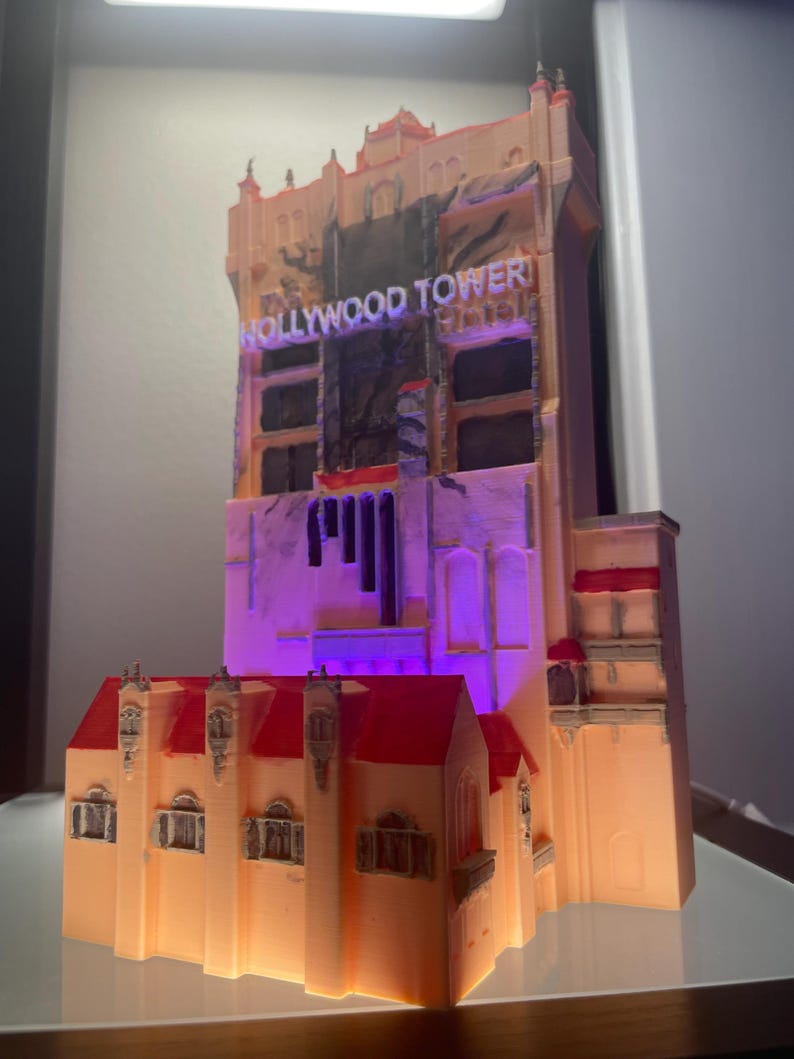 Op de afbeelding: Een 3D-geprint model van het Hollywood Tower Hotel, met een beige buitenkant en rode dakdetails. Het model heeft de verlichte tekst "HOLLYWOOD TOWER". Het gebouw wordt verlicht met paarse en oranje lichten.