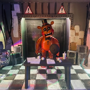 May include: A diorama featuring a brown bear animatronic figure with a black top hat and bowtie standing behind a black desk. The scene includes a chequered floor, a poster with the text CELEBRATE, two warning signs, and a detached animatronic head.